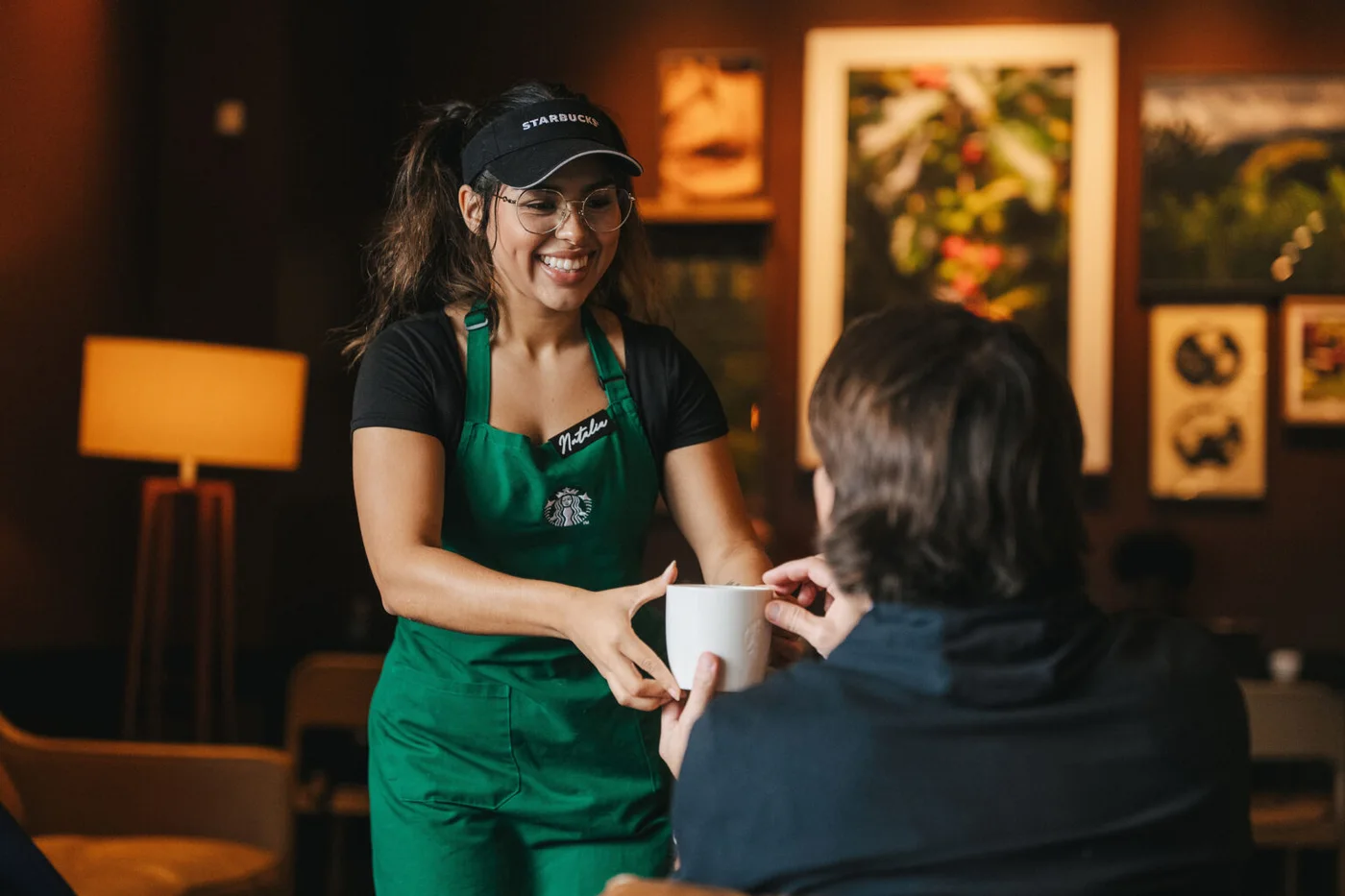 A Starbucks barista in a green apron and cap hands a cup to a customer.